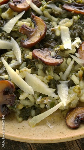 Close-up of creamy gourmet risotto with New Zealand spinach (tetragonia), pan-seared cremini mushrooms, and Parmesan shavings, rotating on a plate. Rustic dark-wood background, vertical video.