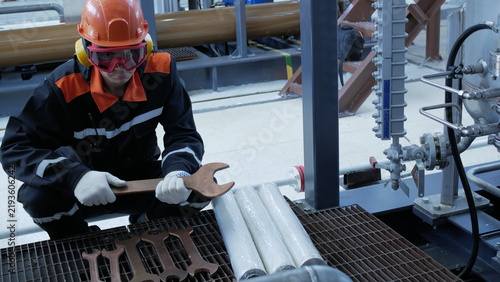Photography Industrial worker crouches with wrench in refinery, wearing hard hat and safety goggles, focused on valve assembly