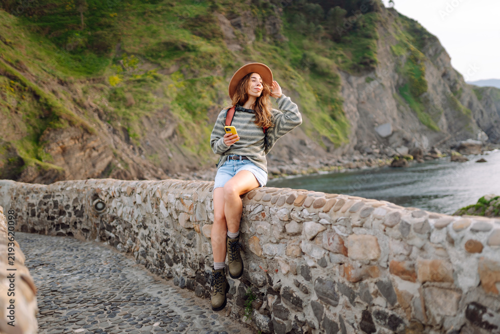 Naklejka premium Woman wearing hat and holding phone stands on stone bridge of fortress overlooking the ocean. Traveler with backpack admires historic architecture. Concept of history, nature, and enjoyment.