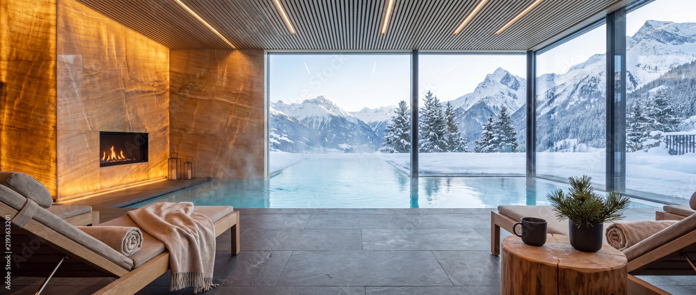 Fototapeta premium Indoor pool overlooking snowy alpine mountains with modern fireplace, warm stone wall, lounge chairs, and panoramic glass windows for luxury winter wellness resort travel
