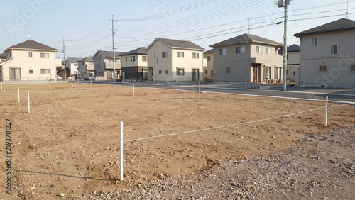 Plakat 白い杭と水糸で区画された分譲住宅地の更地と奥に並ぶ新築住宅を晴天の昼間に写した写真