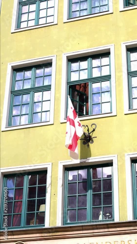 Georgian national flag mounted on building facade in urban setting. White fabric with red cross design hangs prominently against architectural exterior, clearly marking cultural diplomatic or