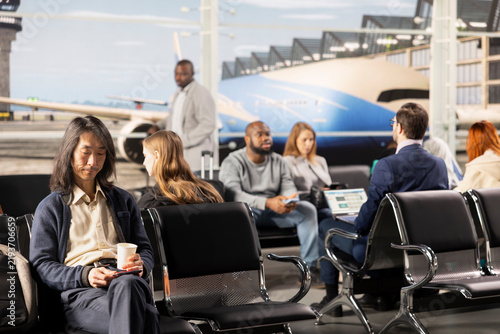 Konstfotografi Bored travelers sit on chairs in a busy airport lounge waiting for departure during a long layover as other passengers prepare to fly
