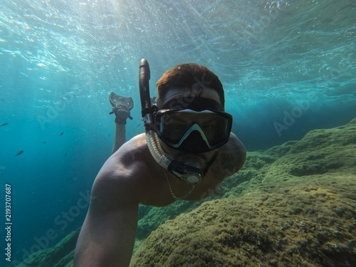 Man snorkeling underwater in mask and fins near rocky seabed in clear blue sea with sun rays breaking surface