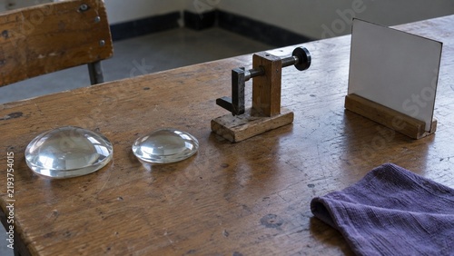 Two glass convex lenses and a wooden optical bench with a screw clamp sit on a worn wooden table beside a white card and a purple cloth