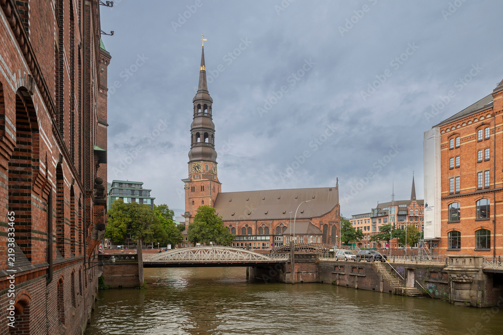 Obraz premium Hamburg, Germany, September 13, 2026 — View along a canal towards St. Catherine's Church with brick buildings and a bridge crossing the water.