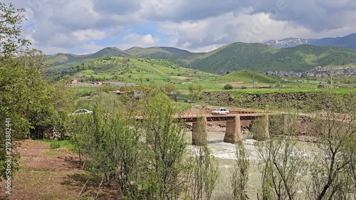 Old stone bridge arches over a swift, foaming river, framed by lush trees, with a distant town and snow-streaked mountains beneath dramatic clouds.
📍 Dorud, Sarvabad, Kurdistan 