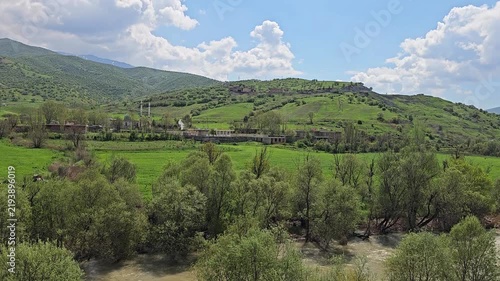 Swift river winds beneath dense green trees, framed by vibrant fields, scattered houses on the hillside, and rolling mountains under a bright blue sky.
📍 Dorud, Sarvabad, Kurdistan 