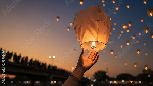 Person releasing a glowing paper lantern into the night sky during a festival with many floating lights