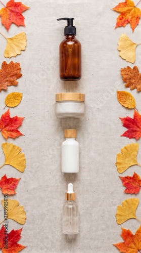 Autumn flat lay composition with organic cosmetic bottles aligned in the center and framed by colorful fall leaves on linen background, seasonal natural skincare concept