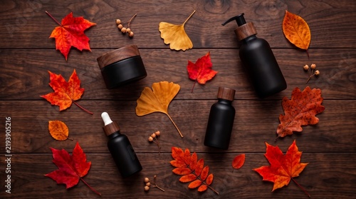Matte black cosmetic bottles and scattered colorful fall leaves on rustic wooden background, premium natural skincare and wellness, flat lay packaging mockup