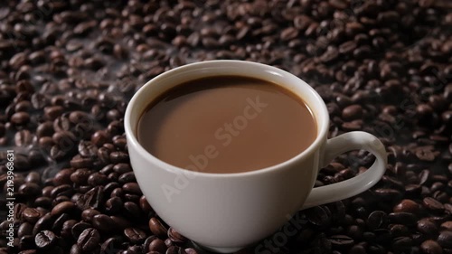 A cup of coffee placed on roasted coffee beans with a slow coffee drop falling into the drink, creating ripples on the surface for food and beverage advertising.