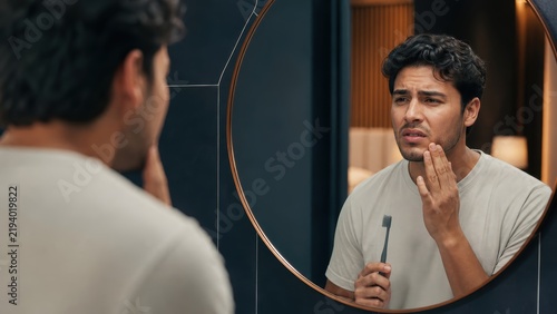 Young man with toothbrush touching sore jaw in bathroom mirror, dental pain concept