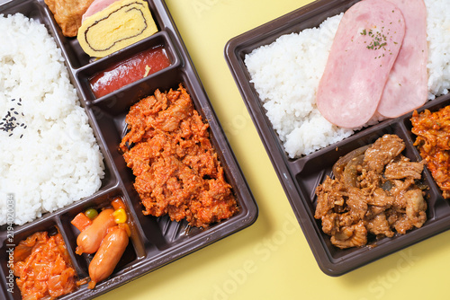 Korean lunch box with rice, meat, and kimchi in plastic packaging on yellow background close-up, top view.