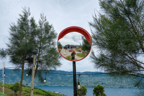 A creative shot of a red circular mirror reflecting a photographer at a scenic viewpoint. Pine trees, string lights, lake, and mountains create a beautiful composition in a tourist spot in Indonesia.