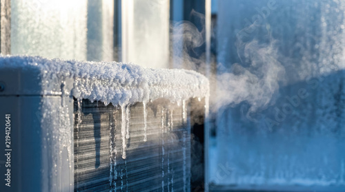 Icy frost melting from heat pump coil outdoors, with dripping icicle and vapor in cold winter sunlight, illustrating frozen equipment and seasonal maintenance needs