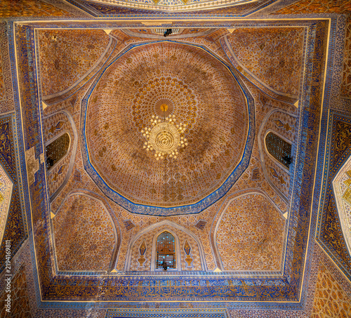 Wide interior view of the main domed chamber at the Amir Temur Mausoleum, also known as Gur-e Amir, in Samarkand. The ceiling is covered with dense gilded floral and geometric ornament, framed by blue