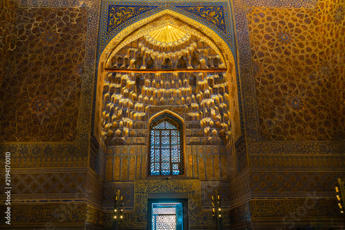 Interior architectural detail from the Amir Temur Mausoleum in Samarkand, showing a recessed arched niche covered with three-dimensional golden muqarnas, blue-and-gold geometric tilework, floral patte