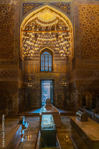 Interior view of the Amir Temur Mausoleum in Samarkand showing stone cenotaphs beneath a richly decorated arched niche. The niche features layered golden muqarnas, blue-and-gold geometric tile work.