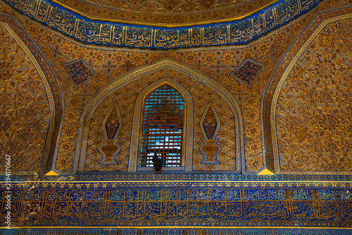 Detailed interior wall of the Amir Temur Mausoleum in Samarkand, featuring a blue-and-gold Arabic calligraphy band, a pointed arched lattice window, symmetrical vase motifs, and dense gilded floral de