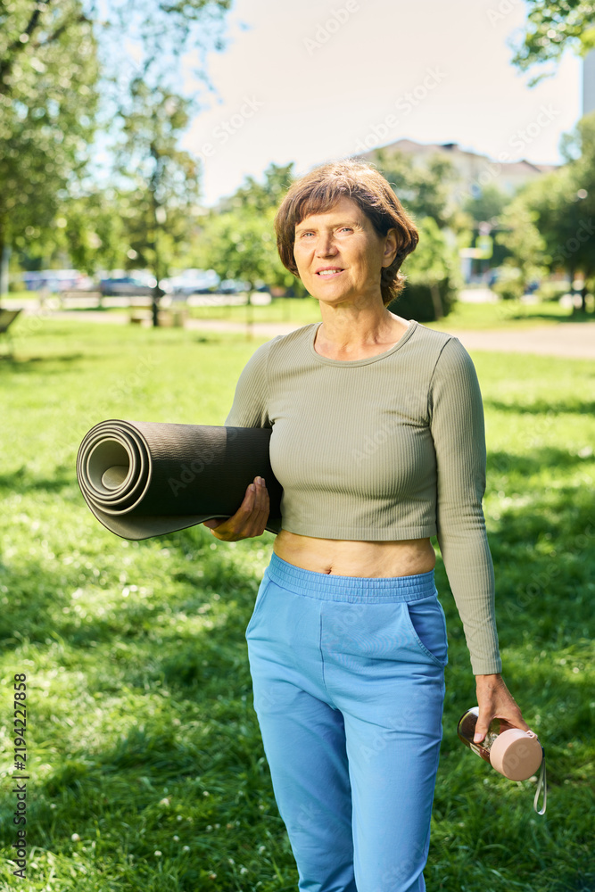Obraz premium Portrait of middle aged Caucasian woman holding rolled yoga mat and water bottle in park. Image ideal for senior fitness campaigns and wellness marketing, vertical