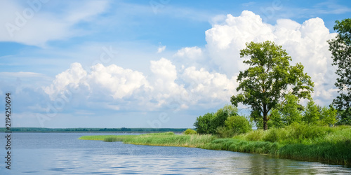 Scenic summer landscape with calm lake, green shoreline, trees and blue sky with clouds, peaceful nature background