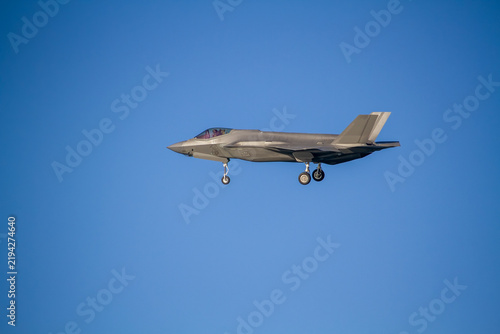 F-35A Lightning II fighter aircraft flying across a blue sky with wheels down, photographed in side profile showing its distinctive stealth aircraft design.