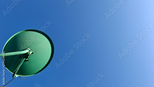 Minimalist bottom view of a green parabolic satellite receiver dish isolated on a vast clear blue sky. Symbol for satellite communication, media transmission, and signal network.