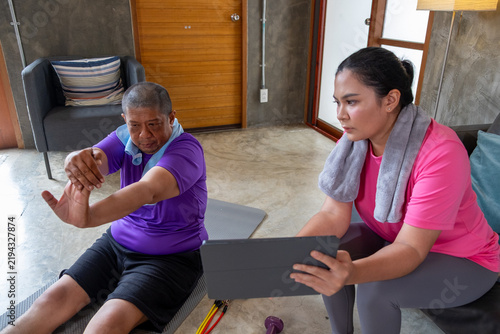 Exercise session shows fat older man stretching beside woman with tablet in bright indoor wellness room, creating supportive moment of focused movement and guidance