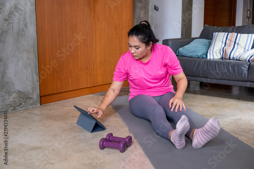 Fat woman doing home exercise while following tablet workout on yoga mat with dumbbell in modern living room