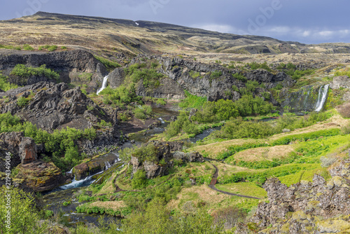 Obraz na plátne Gjain reveals Gjarfoss and a smaller cascade above the winding Rauda River in Thjorsardalur, Iceland, framed by basalt walls, dense shrubs, and a curving footpath in a closer elevated overview