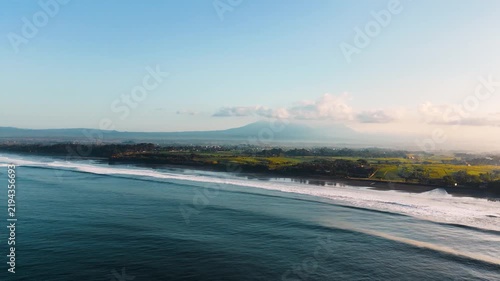 Aerial drone footage of black sand beach coastline with rolling ocean waves, green fields and mountain background at sunrise. Yeh Gangga Tabanan Bali Indonesia scenic tropical landscape Rec709
