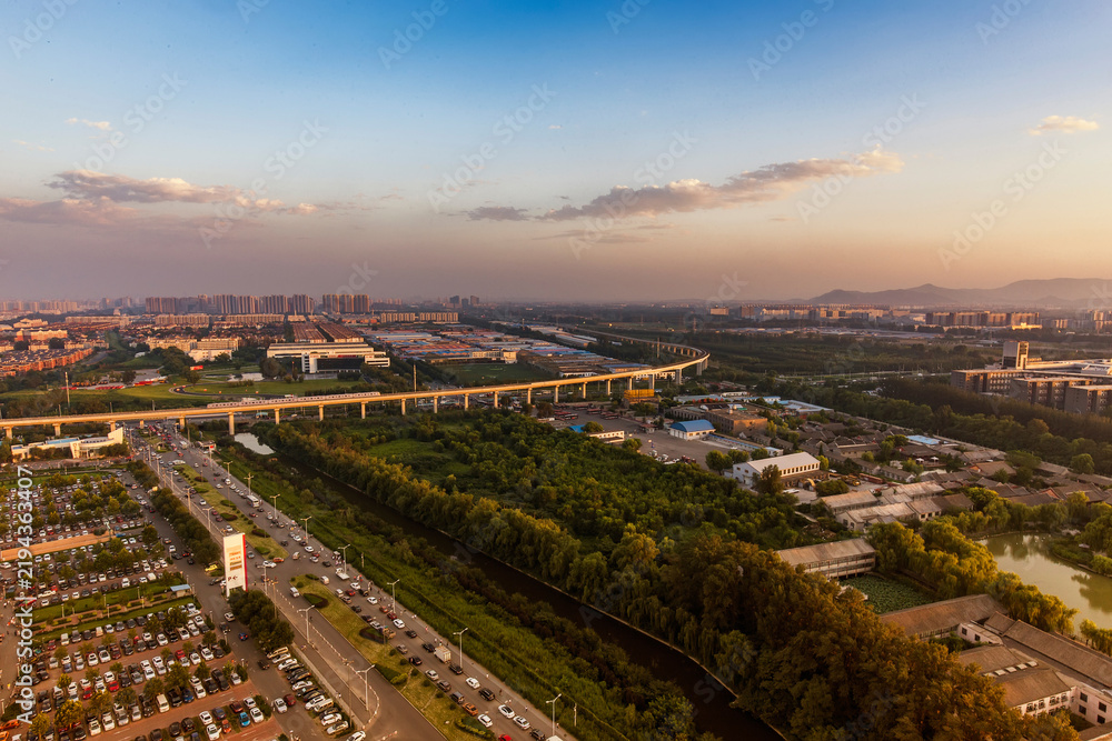 Fototapeta premium Aerial panorama of urban area featuring elevated overpass, roads, green spaces and buildings under sunset sky.