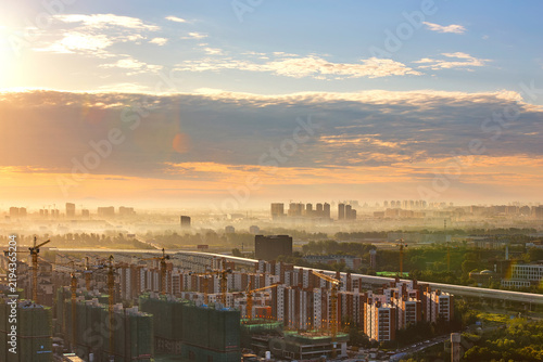 Canvastavla A hazy urban cityscape at sunrise shows ongoing construction with cranes and distant buildings under a colorful sky