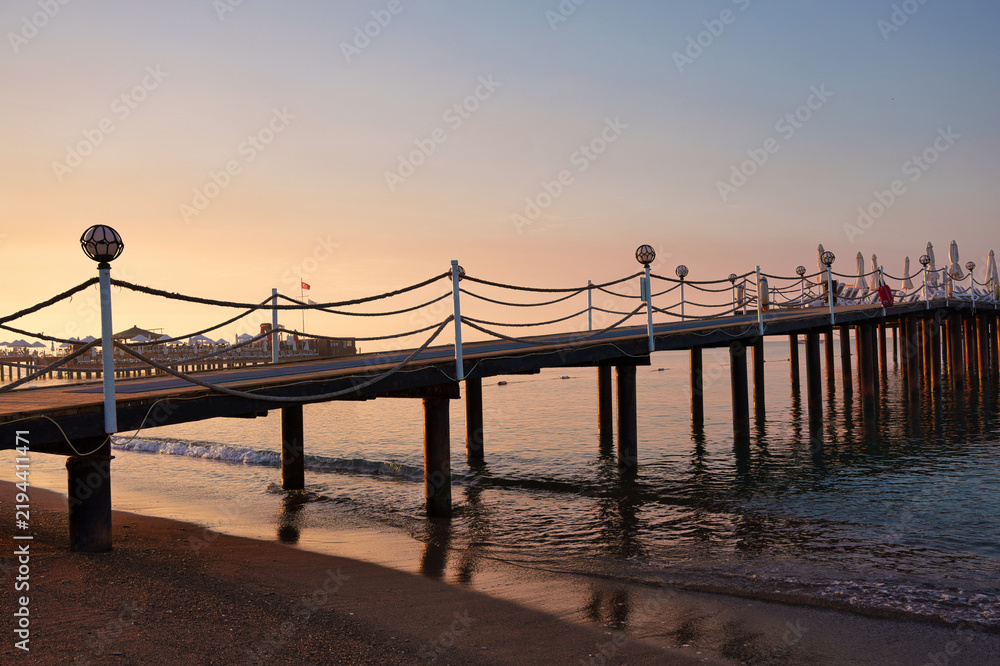 Fototapeta premium Long wooden pier with rope railings on sandy beach during golden sunrise at luxury resort