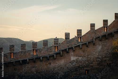 A curving crenellated fortress wall lined with weathered merlons overlooks distant blue mountain ranges beneath a soft cloud-streaked dusk sky, creating a peaceful and atmospheric medieval hilltop sce