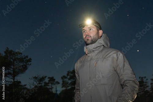 Konstfotografi Man wearing headlamp torch and outdoor jacket standing in forest looking away du