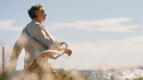 Man walks barefoot through golden grass fields. Wind lifts his shirt as he turns toward distant horizon. Sunlight glows softly through thin clouds above. He appears peaceful, calm