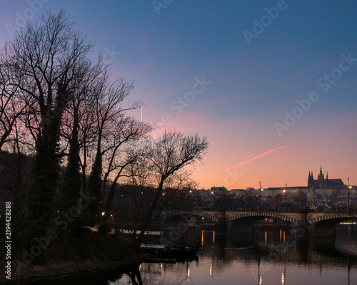 Scenic twilight panorama over Prague castle and river bridge framed by silhouetted winter trees. Serene evening sky with pink and blue gradients casting warm reflective light on calm city waters.