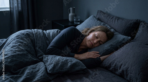 Middle Aged Woman Sleeping Peacefully in Cozy Bed with Dark Linen Bedding, Comfortable Bedroom Interior, Relaxed Female Resting During Night, Serene Sleep Concept, Home Lifestyle, Longevity, Wellness