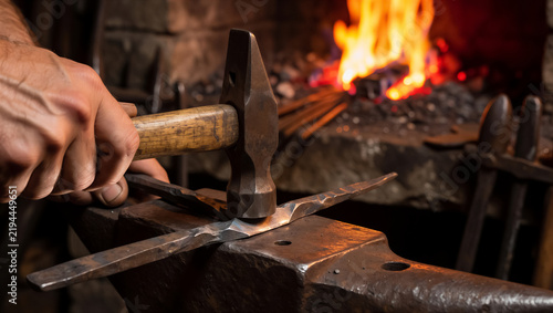 Close-up of a blacksmith striking hot iron on an anvil in a workshop. A bright fire glows in the forge background during the traditional metalwork process