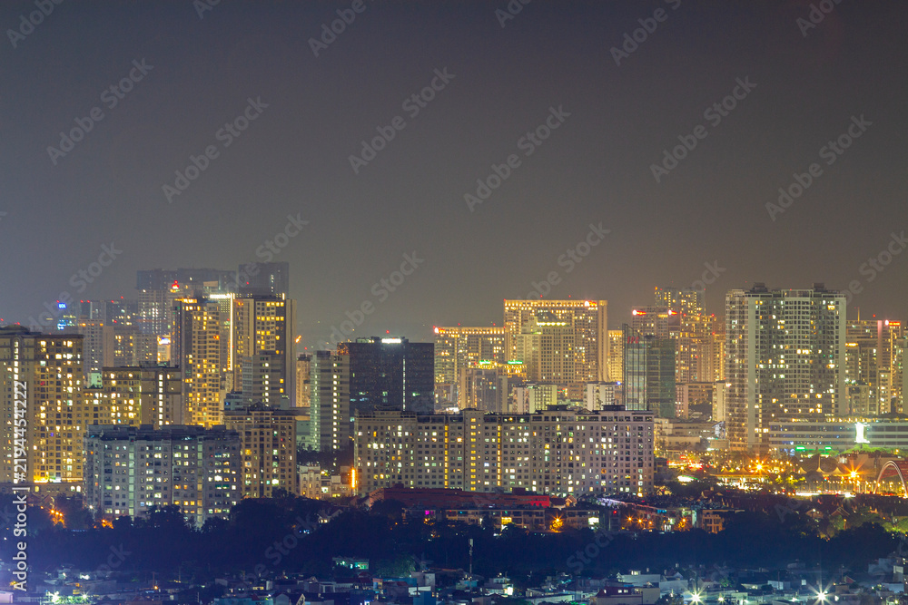 Fototapeta premium A panoramic nighttime cityscape shows dense high-rise buildings, glowing windows, rooftops, and street lights beneath a hazy dark sky with ample copy space.