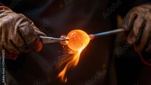 Molten glass artisan shaping glowing amber glass with metal tools in dark workshop