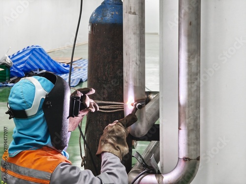 A close-up shot of a professional welder wearing a flip-up protective welding mask and heavy leather gloves, holding a TIG torch and filler rod to perform precise welding on a stainless steel pipe 