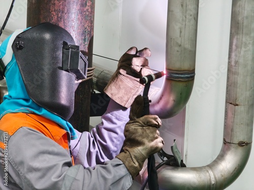 A close-up shot of a professional welder wearing a flip-up protective welding mask and heavy leather gloves, holding a TIG torch and filler rod to perform precise welding on a stainless steel pipe 