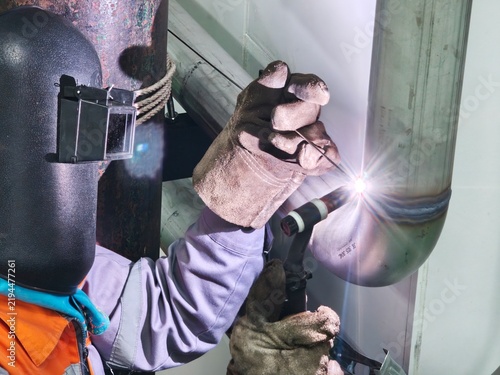 A close-up shot of a professional welder wearing a flip-up protective welding mask and heavy leather gloves, holding a TIG torch and filler rod to perform precise welding on a stainless steel pipe 