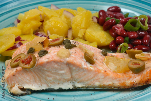 Baked salmon fillet topped with olives capers and lemon served with potato salad and red beans on a blue plate.