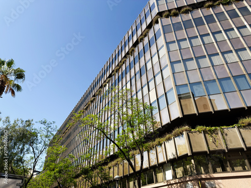 Modern glass office building facade with trees and blue sky in Barcelona, Spain, urban architecture and sustainable greenery