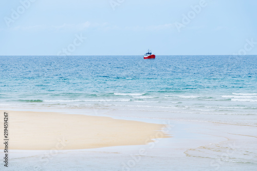 Cuadro en lienzo Red fishing boat offshore beyond sandy island under blue sky