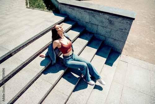 Bild auf Leinwand Young woman relaxing on sunlit stone steps in denim jacket and crop top, enjoyin